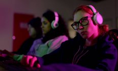 Vista lateral de uma jovem caucasiana gamer trabalhando em um escritório criativo, usando óculos, ouvindo música com fones de ouvido, seus colegas trabalhando em segundo plano — Fotografia de Stock