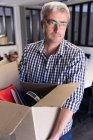 Vista frontal de un hombre caucásico trabajando en una oficina creativa, usando gafas, llevando una caja de cartón . - foto de stock