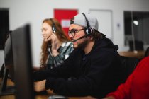Вид збоку молодого кавказького геймера, який працює в творчому офісі, носить окуляри для читання та гарнітуру, дивлячись на екран комп'ютера зі своїм колегою на задньому плані — стокове фото