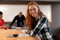 Portrait of a young Caucasian woman sitting at a table in a creative office conference room, smiling to camera with her colleagues at a meeting in the background. — Stock Photo