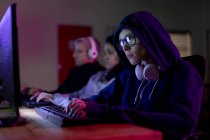 Вид збоку на молоду кавказьку жінку-гравця, що працює в творчому офісі, використовуючи комп'ютер і дивлячись на екран, одягнений в окуляри, капюшон з навушниками навколо шиї і її колеги працюють на задньому плані — стокове фото
