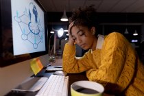 Vista frontal de una joven mujer profesional de raza mixta cansada sentada en un escritorio inclinada su cabeza con los ojos cerrados frente a un monitor de computadora de escritorio - foto de stock