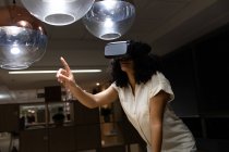Передній вигляд молодої змішаної професійної жінки, яка працює пізно в сучасному офісі, використовуючи гарнітуру VR і вказуючи вгору — стокове фото