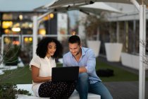 Front view of a young Caucasian professional man and mixed race woman working late at a modern office, sitting on the roof terrace using a laptop computer together — Stock Photo