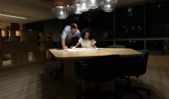 Vista frontal de un joven hombre profesional caucásico y una mujer de raza mixta trabajando hasta tarde en una oficina moderna en un escritorio mirando los planes juntos - foto de stock