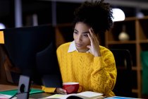 Frontansicht einer jungen Frau mit gemischter Rasse, die spät in einem modernen Büro arbeitet, an einem Schreibtisch sitzt, sich an einen Computerbildschirm lehnt und in der Hand eine Tasse hält. — Stockfoto