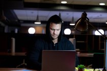 Vista frontal de un joven profesional caucásico que trabaja hasta tarde en una oficina moderna, sentado en un escritorio con una sudadera con capucha usando una computadora portátil - foto de stock