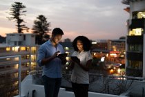 Vista frontale di un giovane professionista caucasico e di una donna mista che lavora fino a tardi in un ufficio moderno, in piedi sulla terrazza sul tetto usando tablet — Foto stock