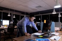 Vista frontal de un joven profesional caucásico que trabaja hasta tarde en una oficina moderna, de pie en un escritorio mirando un monitor de computadora de escritorio - foto de stock