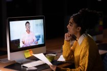 Vista lateral de una joven profesional de raza mixta que trabaja hasta tarde en una oficina moderna, sentada en un escritorio en una reunión en línea con un joven afroamericano visto en su monitor de computadora - foto de stock