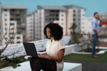 Vue de face d'une jeune femme professionnelle mixte travaillant tard dans un bureau moderne, assise sur la terrasse du toit à l'aide d'un ordinateur portable, son collègue masculin debout en arrière-plan — Photo de stock