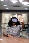 Передній вигляд молодої змішаної професійної жінки, яка працює пізно в сучасному офісі, сидячи за столом в гарнітурі VR, з однією рукою, піднятою перед нею — стокове фото