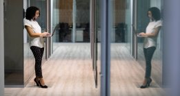 Vista lateral de una joven mujer profesional de raza mixta que trabaja hasta tarde en una oficina moderna, parada en un pasillo usando una computadora portátil, reflejada en una puerta de vidrio - foto de stock