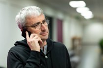Vista frontal de un hombre caucásico con el pelo gris trabajando en una oficina creativa, hablando en el teléfono inteligente, usando gafas - foto de stock