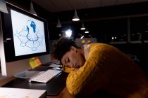Вид збоку на молоду змішану професійну жінку, яка працює пізно в сучасному офісі, спить на столі перед комп'ютерним монітором — стокове фото