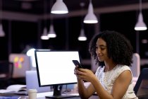 Seitenansicht einer jungen Frau mit gemischter Rasse, die spät in einem modernen Büro arbeitet und mit einem Smartphone am Schreibtisch sitzt — Stockfoto