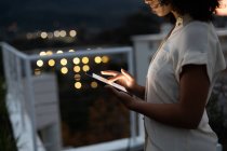 Вид сбоку на молодую женщину смешанного расового происхождения, работающую поздно вечером в современном офисе и стоящую на террасе на крыше с помощью планшетного компьютера — стоковое фото