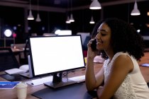 Seitenansicht einer jungen Frau mit gemischter Rasse, die spät in einem modernen Büro arbeitet, am Schreibtisch sitzt und mit dem Smartphone spricht — Stockfoto