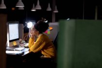 Vista laterale di una giovane donna professionista mista che lavora fino a tardi in un ufficio moderno, seduta su una scrivania appoggiata e fissa un monitor per computer desktop — Foto stock