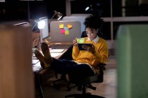 Vista frontal de una joven mujer de raza mixta profesional caucásica que trabaja hasta tarde en una oficina moderna, sentada en un escritorio con los pies en alto usando una tableta y sosteniendo una taza - foto de stock