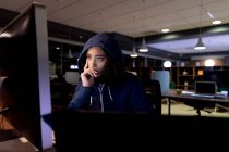 Front view of a young Caucasian professional mixed race woman wearing a hoodie working late in a modern office, sitting at a desk leaning and staring at a desktop computer monitor — Stock Photo