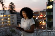 Front view of a young mixed race professional woman working late at a modern office, standing on the roof terrace using a tablet computer — Stock Photo