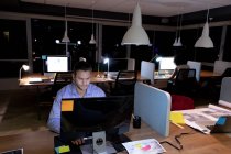 Vista frontal de un joven profesional caucásico que trabaja hasta tarde en una oficina moderna, sentado en un escritorio usando una computadora de escritorio con un colega que trabaja en un escritorio en el fondo - foto de stock