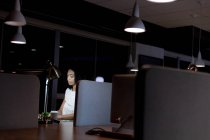 Vista lateral de una joven profesional de raza mixta que trabaja hasta tarde en una oficina moderna, sentada en un escritorio con un café para llevar usando una computadora de escritorio - foto de stock