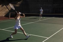 Vista lateral de una joven mujer caucásica y un hombre jugando al tenis en un día soleado, mujer corriendo hacia la pelota - foto de stock