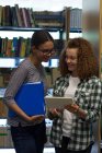 Улыбающаяся девочка-подросток показывает планшетный компьютер другу, стоя в библиотеке — стоковое фото