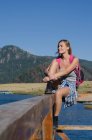 Volle Länge einer lächelnden Wanderin, die auf einem Geländer an der Seebrücke vor strahlend blauem Himmel sitzt — Stockfoto