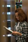 Estudante universitária do sexo feminino lendo livro enquanto em pé pela prateleira na biblioteca — Fotografia de Stock