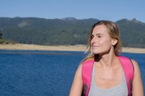 Закройте улыбчивую женщину-туристку, которая смотрит в сторону, стоя на берегу озера — стоковое фото