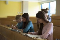 Jóvenes estudiantes universitarios escribiendo en un libro en el escritorio mientras están sentados en clase - foto de stock
