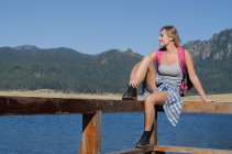 Escursionista femminile sorridente seduta su ringhiera al molo contro il cielo blu — Foto stock