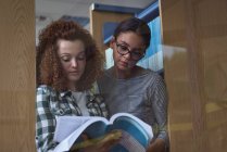 Teenage girls reading book while standing in library seen through glass — Stock Photo