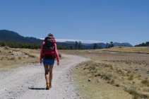 Vue arrière d'une randonneuse avec sac à dos marchant sur un chemin de terre contre le ciel bleu — Photo de stock