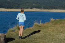 Rückansicht einer Wanderin, die an einem sonnigen Tag auf einem Feld am See steht — Stockfoto