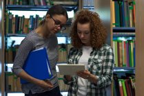 Teenage girl showing tablet computer to friend while standing in library — Stock Photo