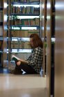 Vista laterale di adolescente lettura libro mentre seduto vicino allo scaffale in biblioteca — Foto stock