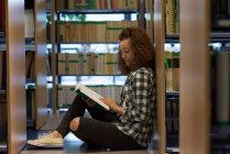 Vista laterale del libro di lettura studentessa adolescente mentre seduta vicino allo scaffale in biblioteca — Foto stock