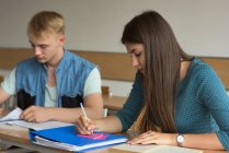 Estudante feminina escrevendo em nota adesiva enquanto estudava na mesa em sala de aula — Fotografia de Stock