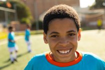 Porträt eines Fußballspielers mit gemischter Rasse, der einen blauen Mannschaftsstreifen trägt, an einem sonnigen Tag auf einem Spielfeld steht, in die Kamera blickt und lächelt, während Teamkollegen im Hintergrund stehen — Stockfoto