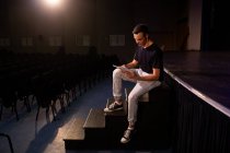 Vista lateral de un adolescente caucásico en un teatro de secundaria vacío, sentado en los escalones del frente del escenario, preparándose para una actuación, sosteniendo un guion y líneas de aprendizaje - foto de stock