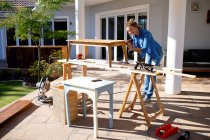 Caucasian woman spending time at home self isolating and social distancing in quarantine lockdown during coronavirus covid 19 epidemic, doing DIY in her garden, drilling wooden table. — Stock Photo