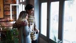 Vista lateral de um jovem mestiço e uma jovem caucasiana desfrutando do tempo em casa, em pé à janela, abraçando e bebendo café. — Fotografia de Stock