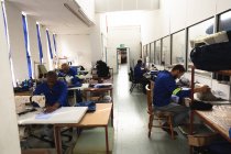 Grupo multiétnico de trabajadores masculinos en un taller en una fábrica que fabrica sillas de ruedas, sentados en un banco de trabajo, utilizando máquinas de coser - foto de stock
