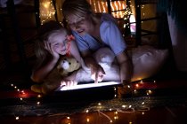 Vista frontal de una mujer caucásica disfrutando de tiempo en familia con su hija en casa juntos, acostada en una tienda de campaña en la sala de estar sonriendo, usando una tableta digital, con su hija abrazando su oso de peluche - foto de stock