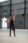 Вид спереди на юношескую гимнастку смешанной расы, выступающую в тренажерном зале, упражняющуюся с обручем, опирающуюся на руки, обруч на ногу, в черном трико — стоковое фото