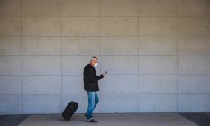 Uomo anziano caucasico in giro per le strade della città durante il giorno, indossando una maschera contro il coronavirus, covid 19, utilizzando uno smartphone e tirando una valigia. — Foto stock